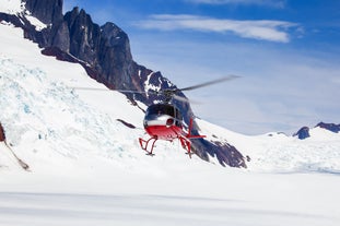 A red and white helicopter flies over a snowy glacier near rugged mountains under a clear blue sky.