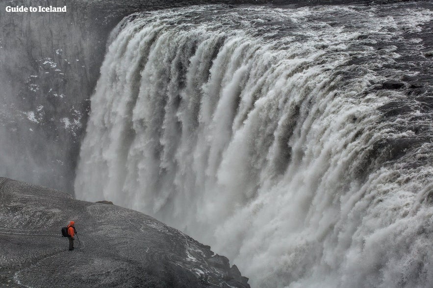 Traveler in orange jacket standing near Dettifoss waterfall in Iceland, Europe’s most powerful waterfall with dramatic mist and flow